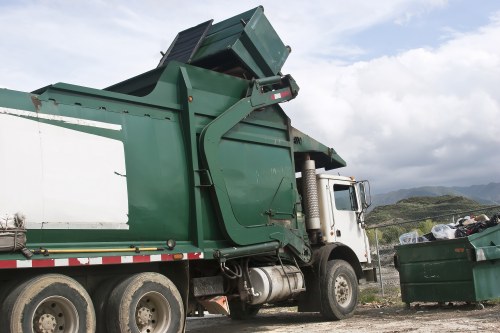A commercial green waste disposal skip mounted on the chassis of a white refuse collection truck, situated outdoors on a gravel surface with a partly cloudy sky overhead. The skip is in a tilted position, indicating it may be in the process of being emptied or loaded, with the upper section visible and open at the top. The truck's cab and side mirror can be seen towards the front, with a large silver exhaust pipe and mechanical components adjacent to the chassis. In the background, there are distant hills and a landscape typical of the Kingston upon Thames area, with some scattered rubbish and a green wheelie bin nearby. The scene reflects a professional rubbish removal operation, likely carried out by Kingston upon Thames House Clearance or a similar service, involving outdoor waste collection and disposal activities.