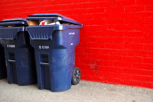 Two large dark blue wheelie bins with black lids positioned on a concrete pavement against a bright red brick wall. The bins have visible scratch marks and weathered surfaces, indicating regular use. The bin on the left has a partially visible white and red sticker, while the right bin shows a wheel at the back corner. The area appears to be an outdoor space, possibly a side alley or driveway, with minimal environmental context. The lighting suggests natural daylight, casting subtle shadows on the ground. The image is associated with Kingston upon Thames House Clearance, highlighting waste collection and rubbish removal services for residential properties. This scene reflects typical rubbish disposal equipment used by local waste management providers in the Surrey area, including Kingston upon Thames.