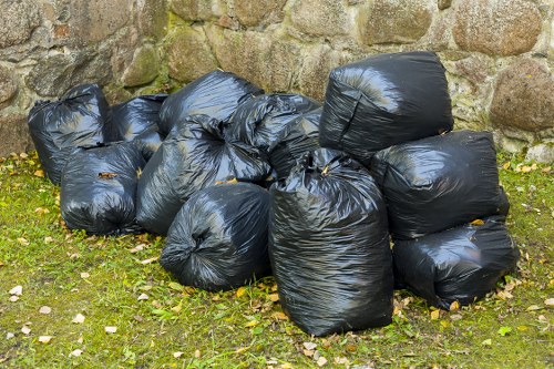 A collection of black plastic rubbish bags filled with waste, stacked loosely on a grassy area near a stone wall. The bags are shiny, slightly bulging, and appear to be tied at the top, with some partially leaning against each other. Small scattered leaves surround the bags on the ground, which is covered in green grass and patches of moss. The stone wall behind the bags is made up of irregularly shaped rocks with a natural, weathered appearance, creating a rural or residential environment that might require rubbish removal services in Kingston upon Thames or the surrounding area. The scene is outdoors with natural lighting, illustrating typical waste collection in a domestic or garden clearance context.