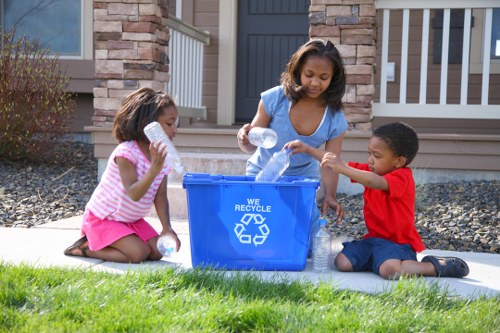 Three children with light to medium skin tones are engaged in recycling activities outdoors in front of a residential property. The girl in the middle, wearing a blue dress, is placing empty plastic bottles into a bright blue recycling bin marked with a white recycling symbol and the words 'WE RECYCLE.' To her right, a boy dressed in a red T-shirt and dark shorts is handing her a plastic bottle, while on her left, another girl in a pink striped top and pink skirt is holding an empty plastic bottle close to her mouth, seemingly preparing to place it into the bin. The scene takes place on a well-maintained lawn, with a paved area beneath the children, and the background features a house with a stone and brick facade, a dark grey front door, and porch railings. The overall environment appears clean and orderly, emphasizing environmentally conscious waste management and recycling habits typical in residential areas near Kingston upon Thames.